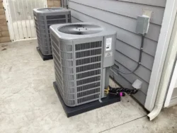 Two newly installed HVAC condenser units mounted on concrete pads beside a gray house exterior, with electrical and refrigerant lines connected.